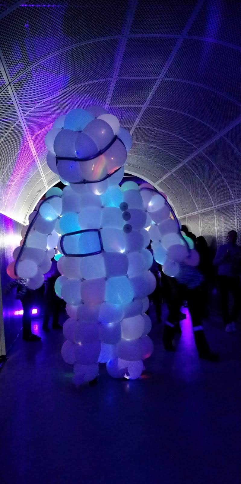 White giant balloon astronaut figure with LED lighting walking through a tunnel entrance at an unknown venue (2019) — Titans of Air